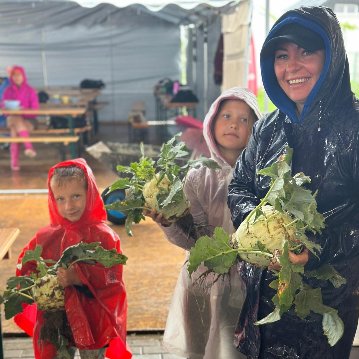 Eine Gruppe von Kindern in Regenmänteln hält zusammen mit einer lächelnden Betreuerin riesige Kohlrabi während einer Aktivität im Freien.