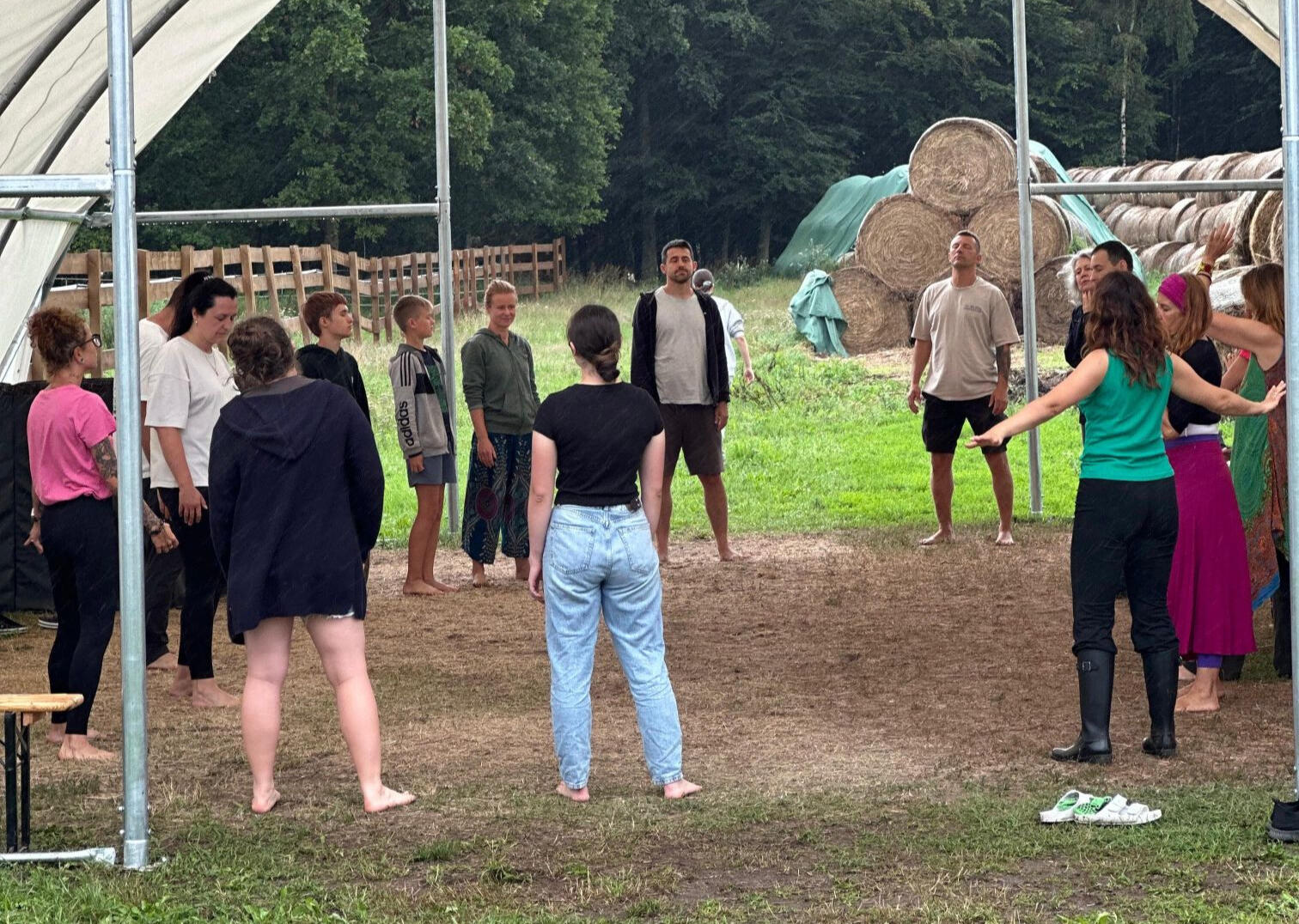 Grupa uczestników warsztatów stoi w kręgu na świeżym powietrzu w Modlimowo Forest Camp, w tle bele słomy i las.