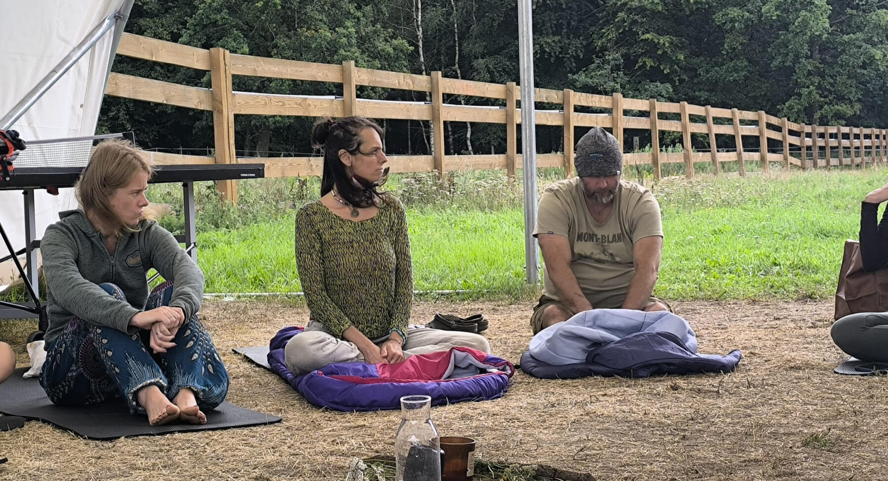 Wyciszenie i dotyk kwantowy w naturze. Uczestnicy siedzący na matach podczas sesji terapeutycznej na świeżym powietrzu, w tle drewniane ogrodzenie i las w Modlimowie.