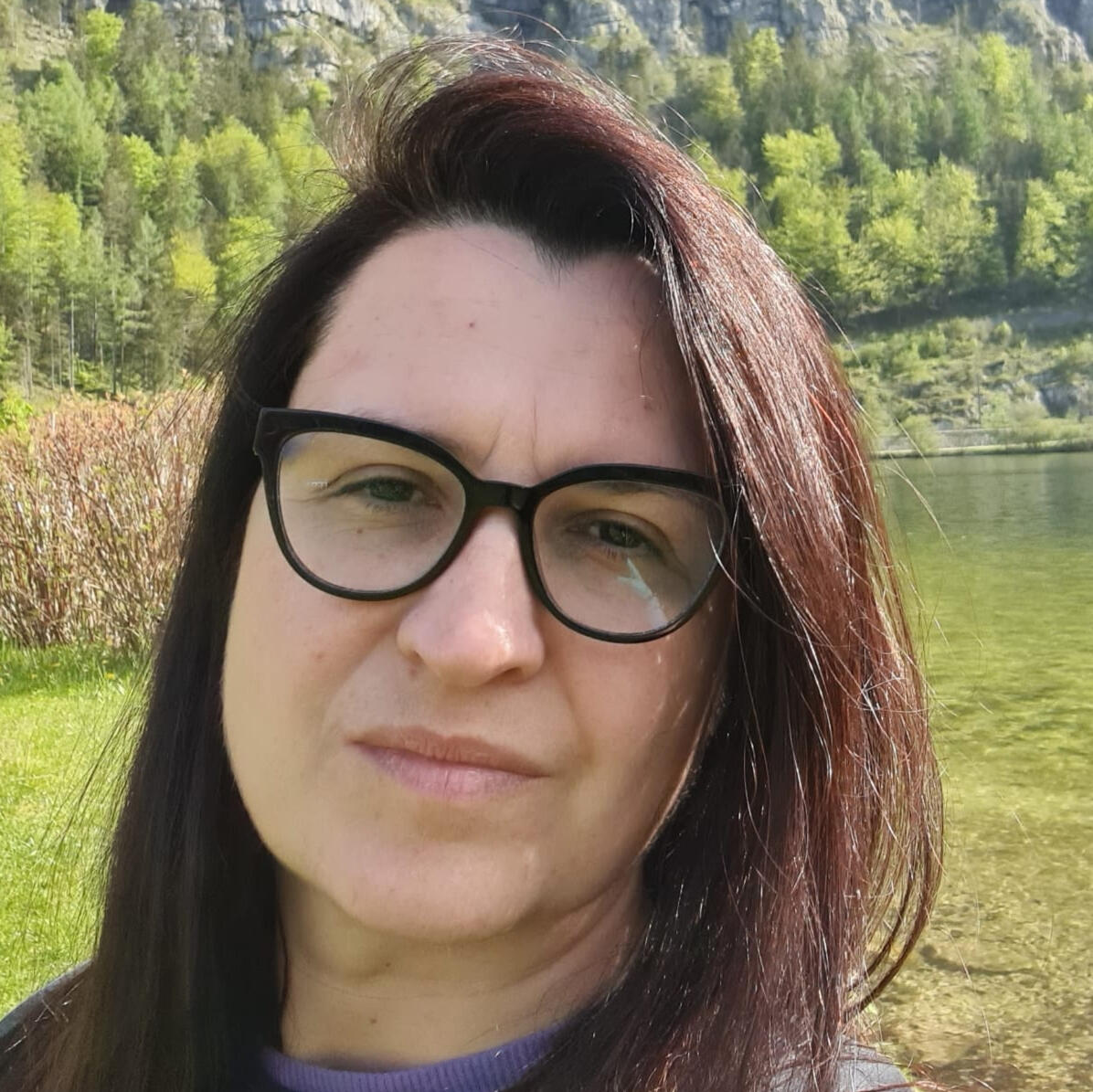 Portret uśmiechniętej prowadzącej w ciemnych okularach na tle jeziora i zielonych wzgórz.